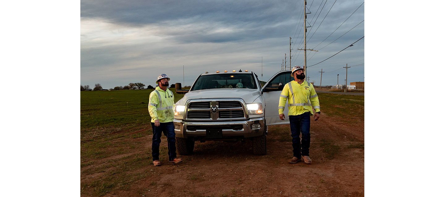 Two electrical linemen in reflective safety gear, Dylan Edgmon and Kevin Walker, stand beside a white work truck with headlights on, parked on a dirt road near power lines.